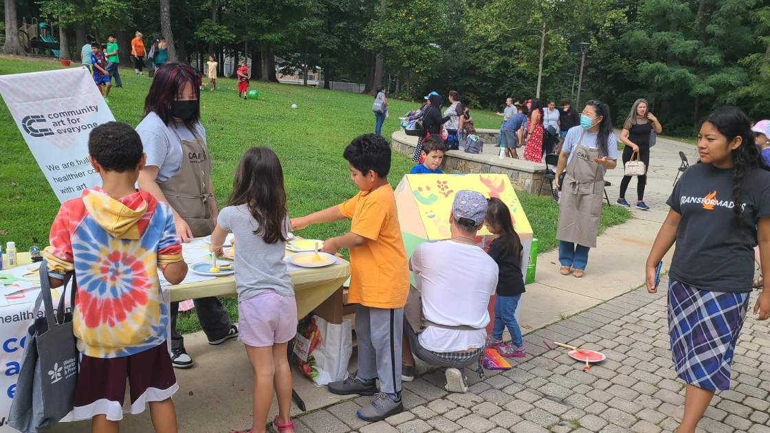 A group of children painting a colorful cardboard box house with the help of CAFE volunteers.